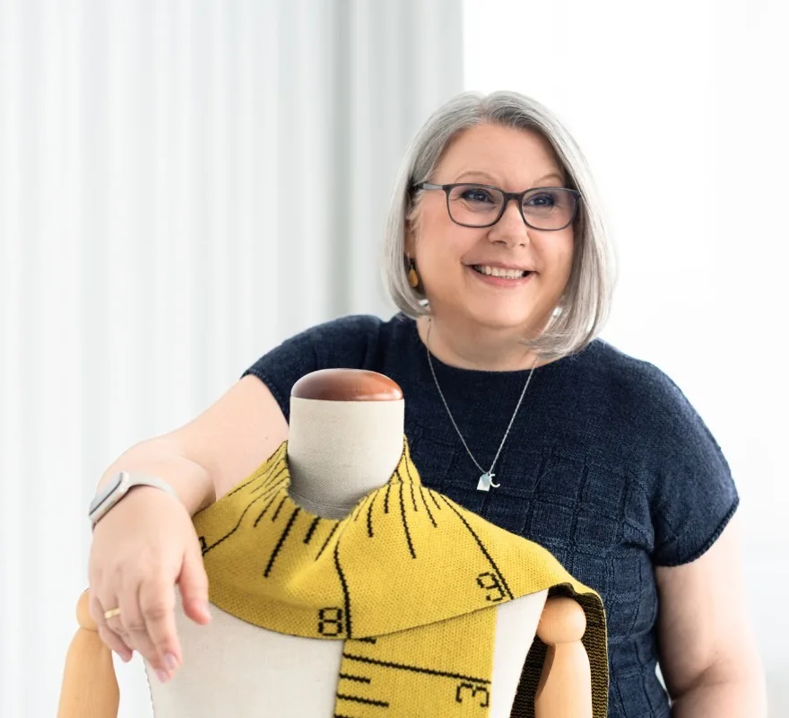 Claudia Eisenkolb lehnt lachend an einer Schneiderpuppe, über der ein gelbes Strickteil mit Maßband-Muster liegt
